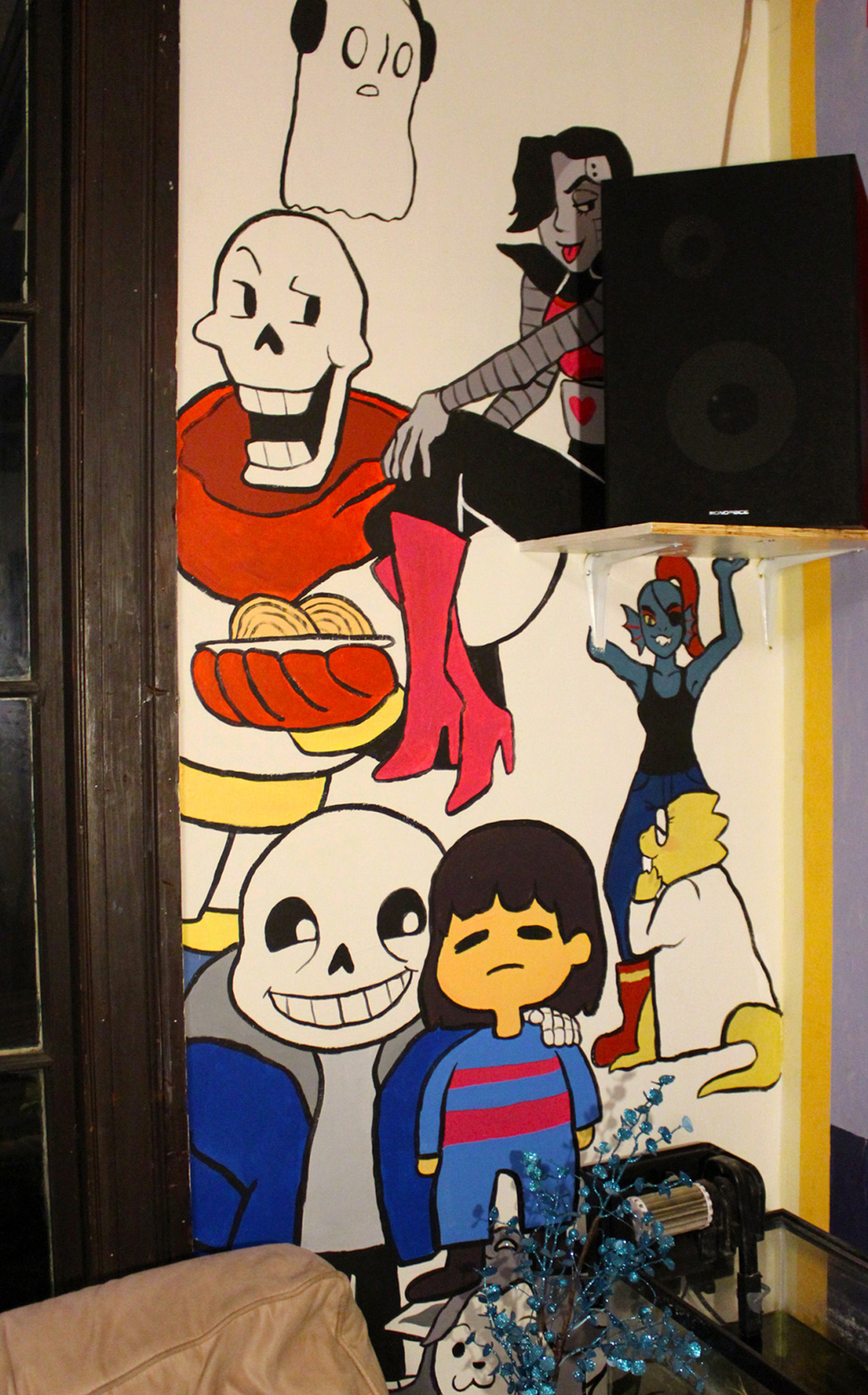 A mural of several Undertale characters: Napstablook, Mettaton, Papyrus, Sans, Frisk, Undyne, and Alphys. The way it is constructed in the room it looks like Undyne is holding up a shelf that Mettaton is sitting on.